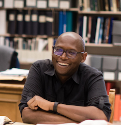 Julian sitting at a table with arms crossed, smiling, with books in the background