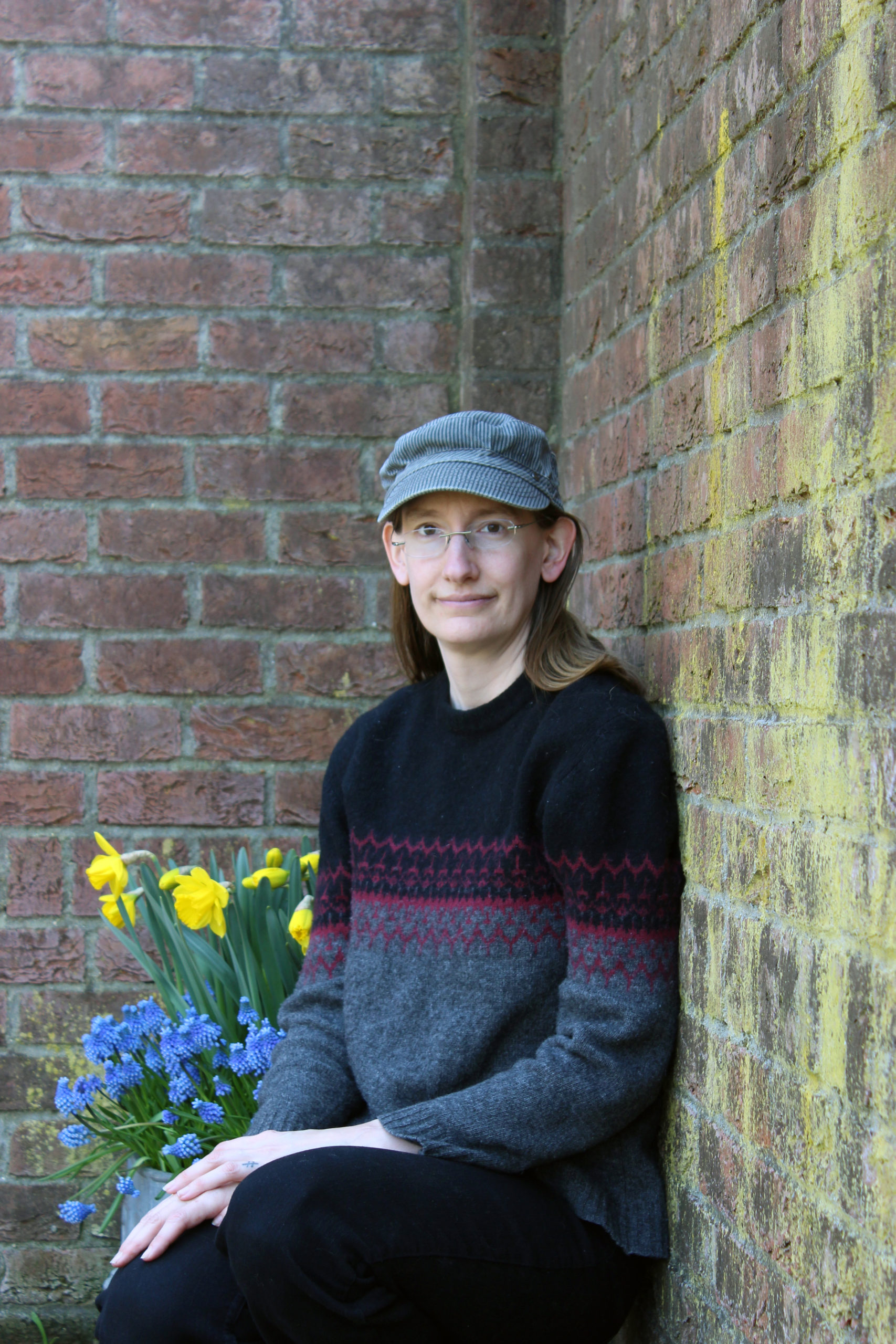 Headshot of Amanda Ticker sitting and looking forward, wearing glasses and a hat with a brick background