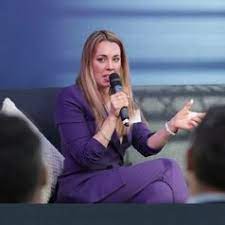 Rachel Adams, seated crosslegged on a couch with pillows, speaks into a microphone and gestures with her left hand