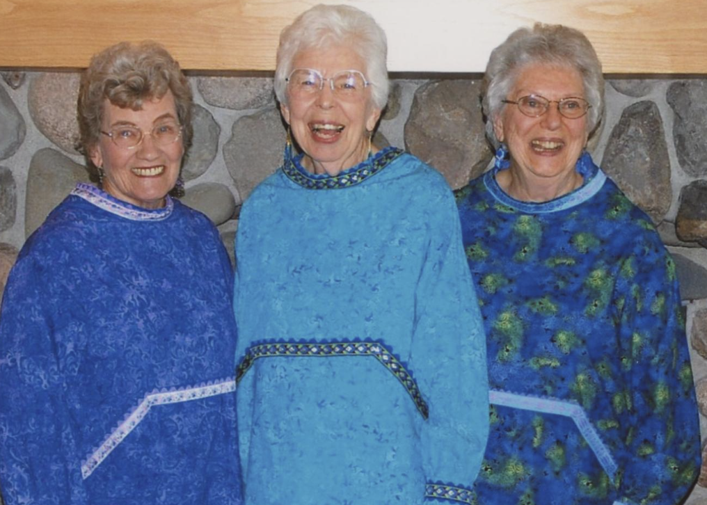 In the image, Anne (left), Jacque (center), and Marge (right) pose for a photo in 2010. They are wearing traditional Alaska Native Kuspuk attire.